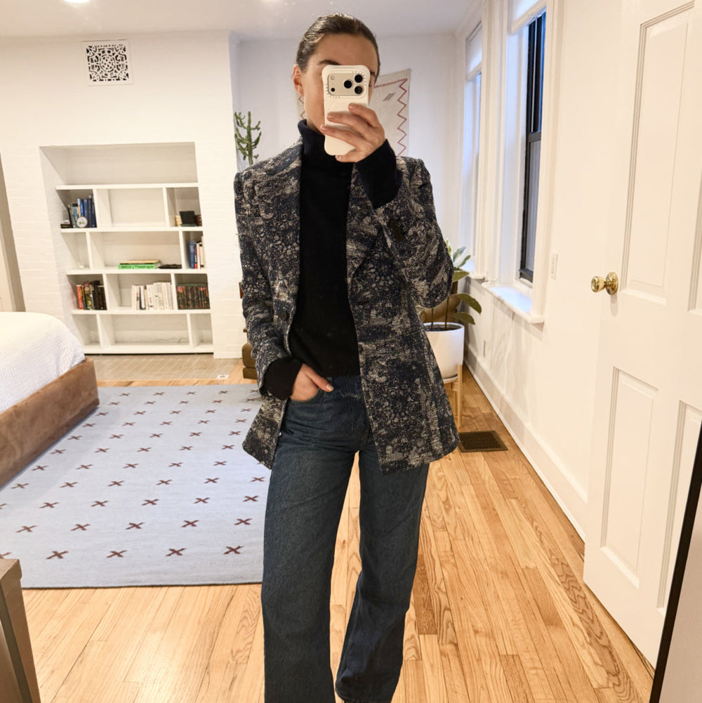 Person taking a mirror selfie wearing a patterned coat, black shirt, and jeans in a bedroom.