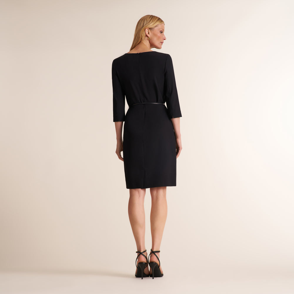 A woman modeling a black knee-length dress with three-quarter sleeves and a thin waist tie, shown from the back against a plain beige studio background.