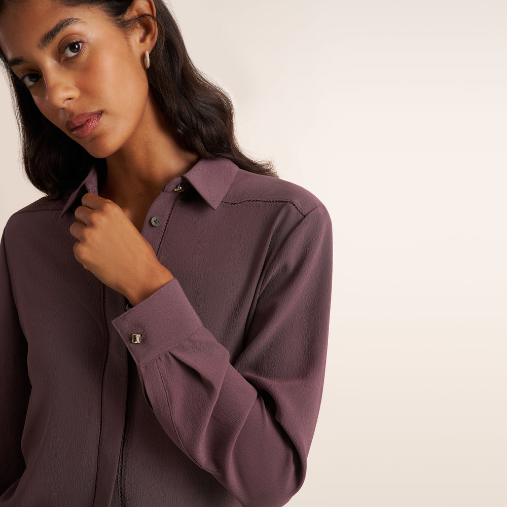 A woman wearing a long-sleeve button-down shirt in a muted mauve-purple color, shown in a close-up shot against a plain off-white background.