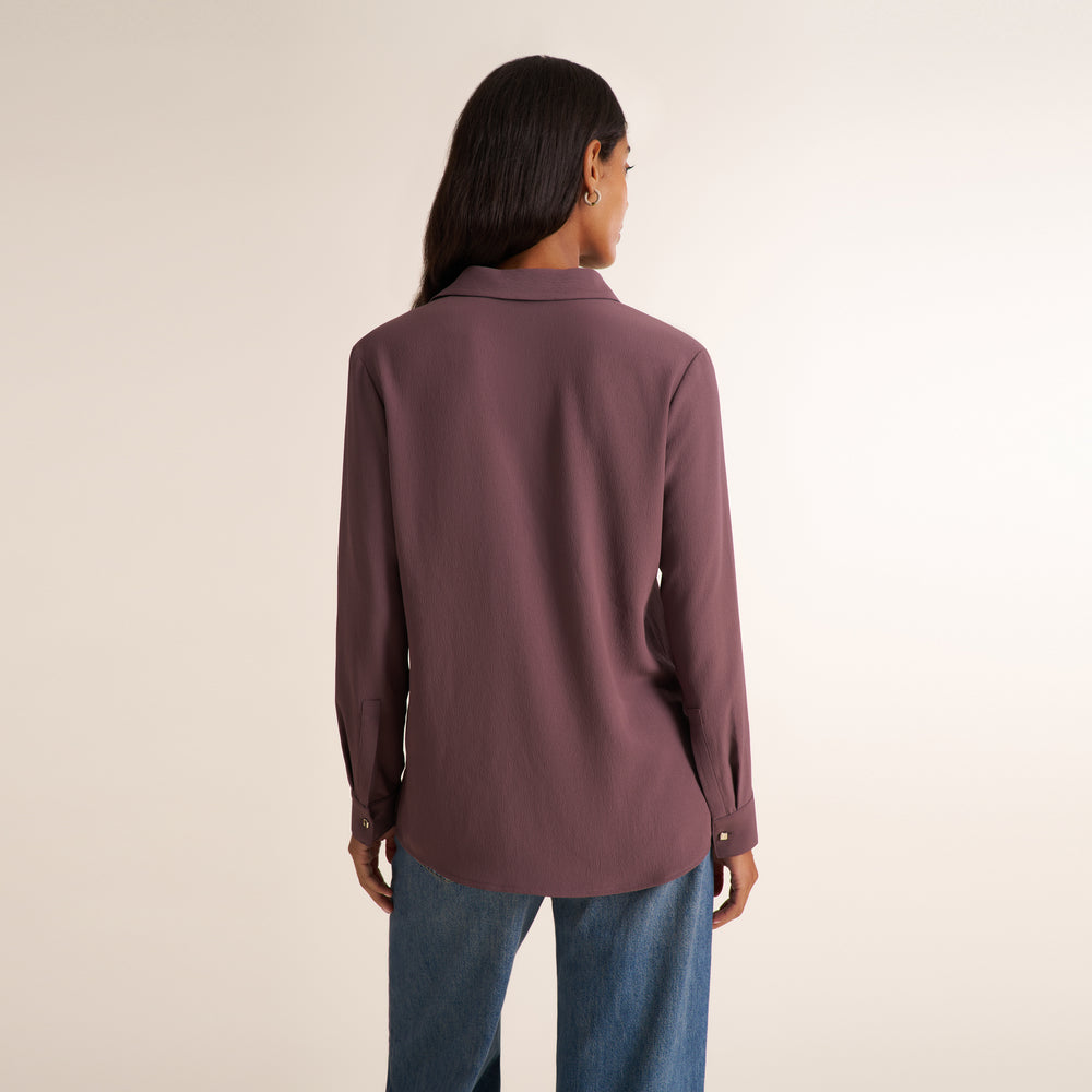 A woman viewed from the back wearing a long-sleeve collared blouse in a muted dusty mauve color, paired with blue jeans, against a plain light beige studio background.