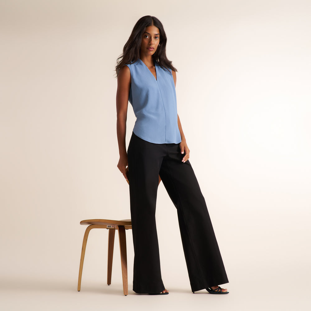 A woman models a sleeveless light blue blouse and black wide-leg pants, standing next to a wooden stool against a warm beige studio background.