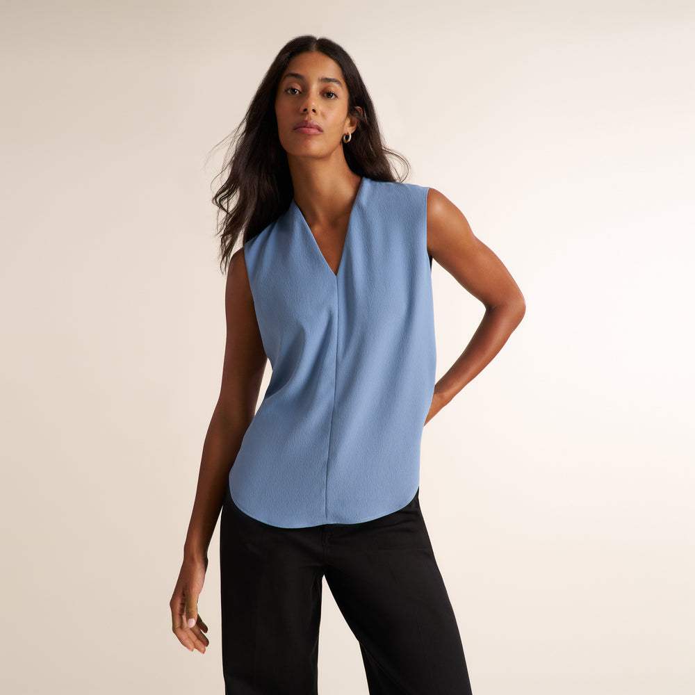 A woman modeling a sleeveless light blue wrap-style blouse with a V-neckline, paired with black pants, against a plain beige studio background.