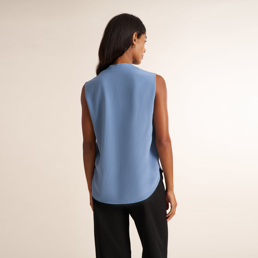 A woman shown from the back wearing a sleeveless light blue top and black pants, standing against a plain light beige studio background.