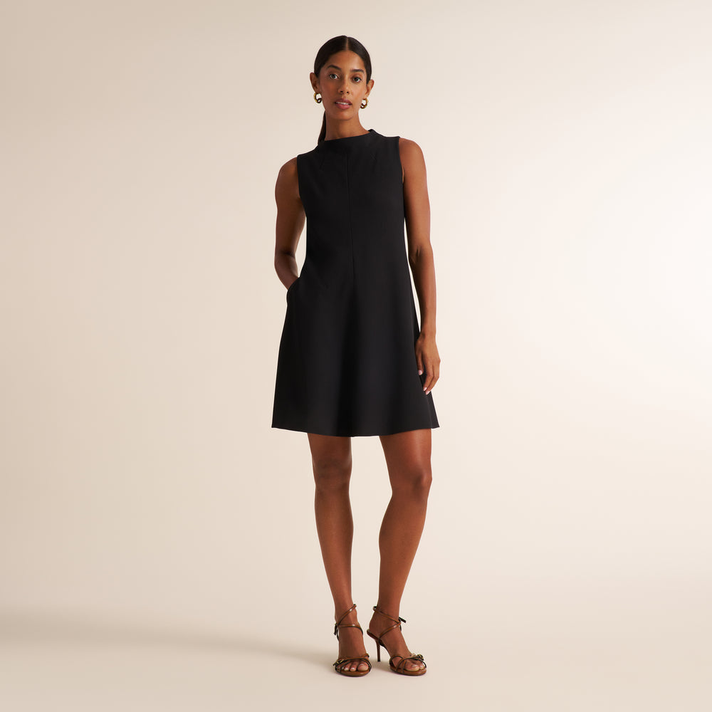 A woman modeling a sleeveless black A-line mini dress with a high mock neck and side pockets, paired with brown strappy heels, against a solid beige studio background.
