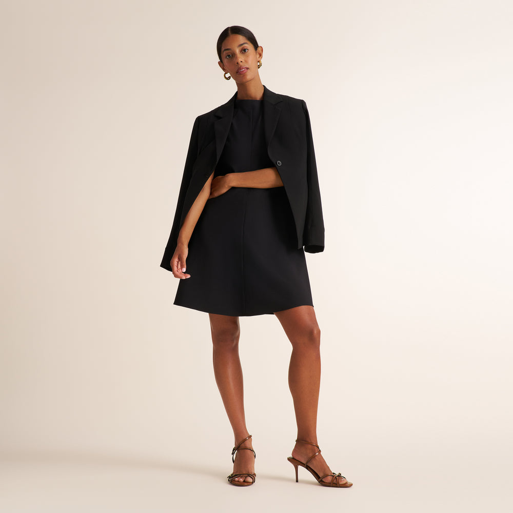 A model poses in a black cropped blazer and matching black mini skirt, styled with a black top and brown strappy high-heeled sandals against a solid light beige studio background.