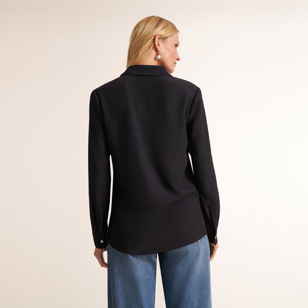 A woman shown from the back wearing a black long-sleeve collared blouse and blue jeans against a plain light beige studio background.