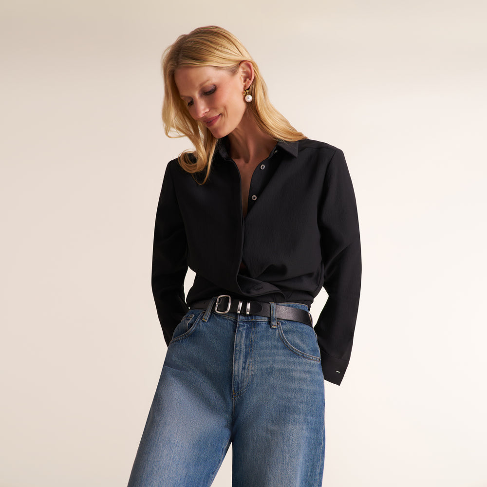 A woman wearing a black long-sleeve button-up shirt tucked into blue jeans with a black belt, photographed against a plain light beige studio background.