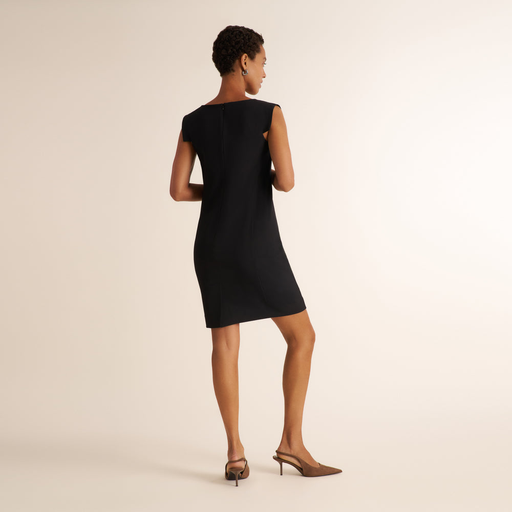A woman modeling a black sleeveless fit-and-flare dress from the back, paired with brown pointed-toe slingback heels, against a solid warm off-white studio background.
