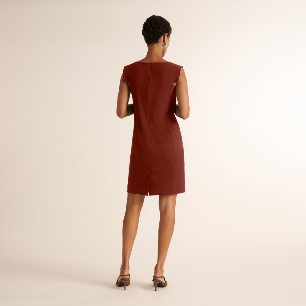 A woman viewed from the back wearing a sleeveless, knee-length rust-brown dress and dark heeled sandals against a plain warm off-white studio background.