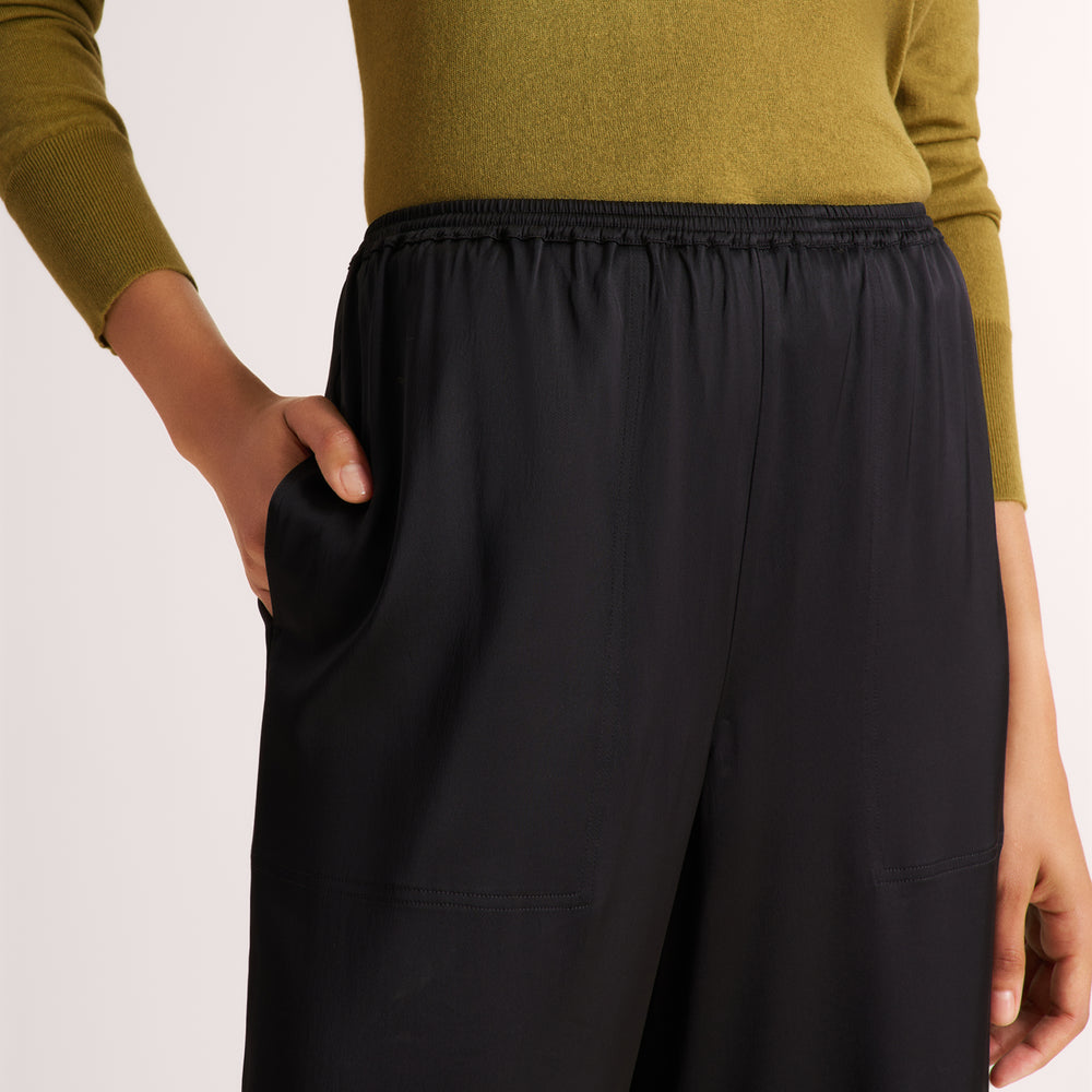 A close-up of a person wearing black wide-leg pants with an elastic waistband and side pockets, paired with an olive green long-sleeve top against a light neutral background.