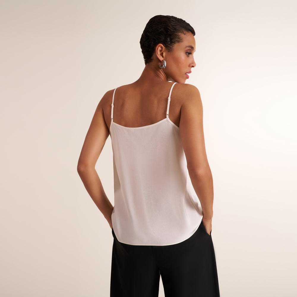A woman shown from the back wearing a light cream/off-white spaghetti-strap camisole top and black pants, standing against a plain light beige studio background.