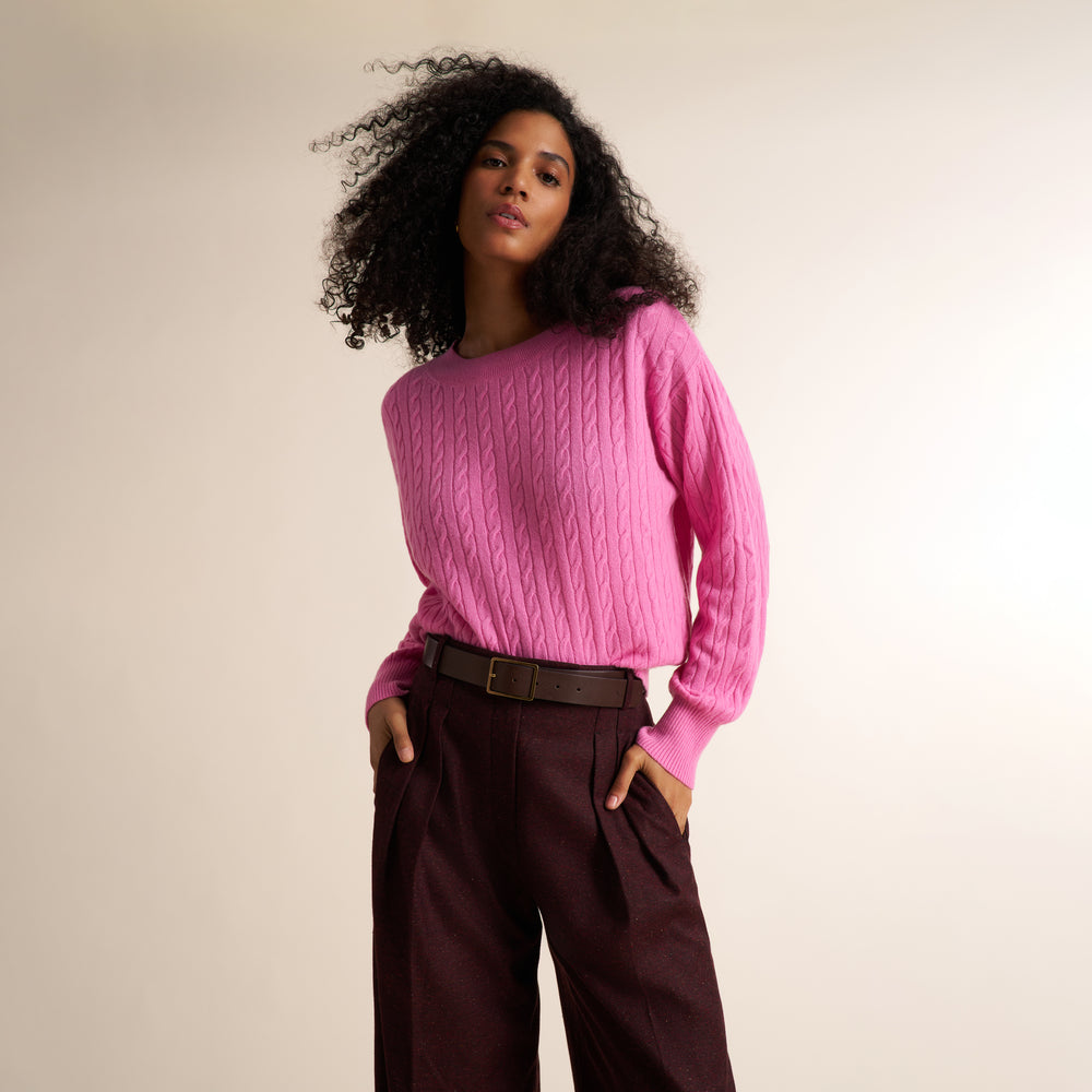 A woman with curly black hair wearing a bright pink cable-knit sweater and dark brown pleated trousers, standing against a plain light beige studio background.