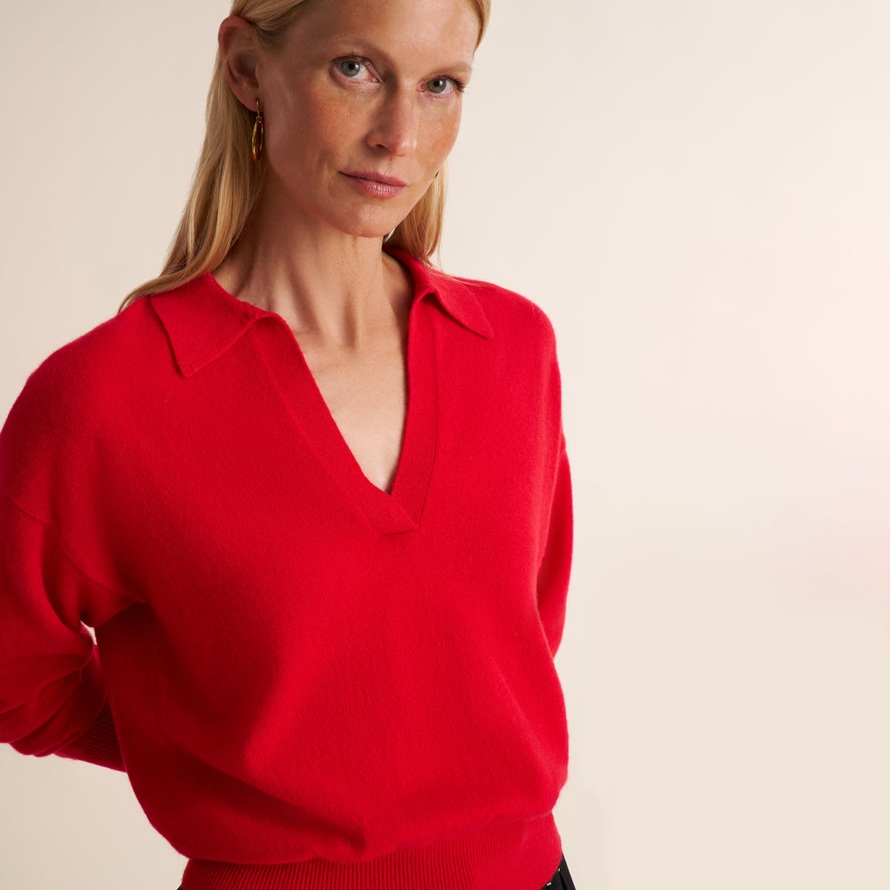 A woman wearing a bright red knit polo-style sweater with a V-neck collar, photographed against a plain light beige studio background.