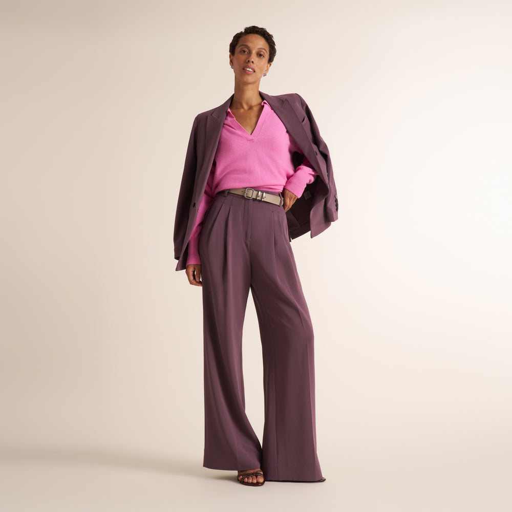 A brunette woman modeling a pink long-sleeve knit top with a split V-neck collar, paired with purple trousers and purple blazer over her shoulders, against a plain light beige studio background.