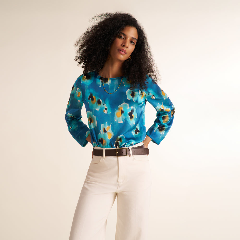A woman wearing a long-sleeve satin blouse with a blue, teal, black, and gold abstract pattern, paired with white trousers and a dark brown belt, against a plain off-white background.
