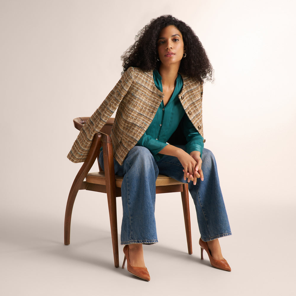 A woman sits on a wooden chair wearing a beige and brown plaid blazer over a teal blouse, blue jeans, and brown heels against a plain light beige studio background.