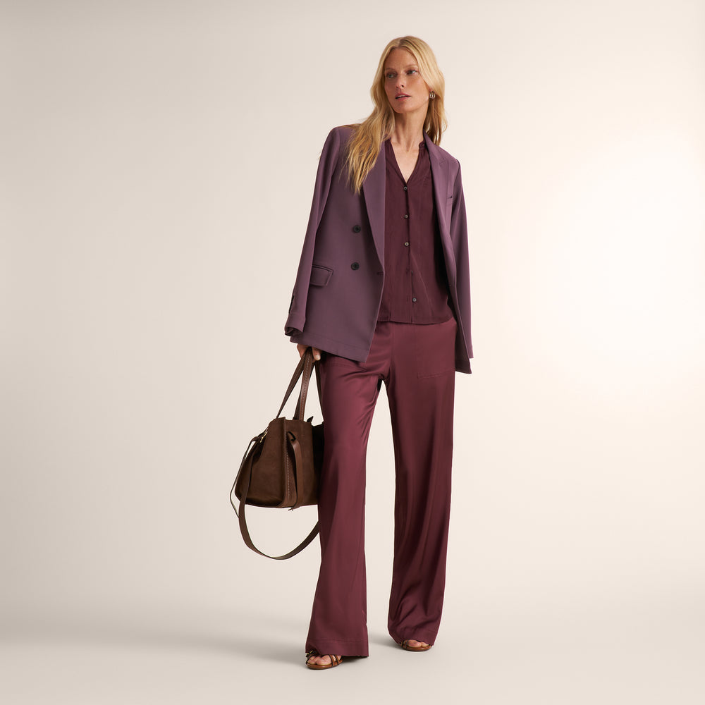 A woman models a matching mauve-purple double-breasted blazer and wide-leg trousers set with a matching button-up shirt, holding a brown leather handbag against a plain beige studio background.
