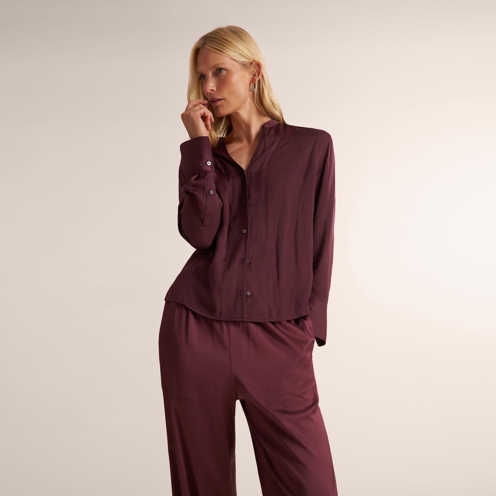 A woman modeling a matching burgundy button-up shirt and trousers set against a plain light beige studio background.