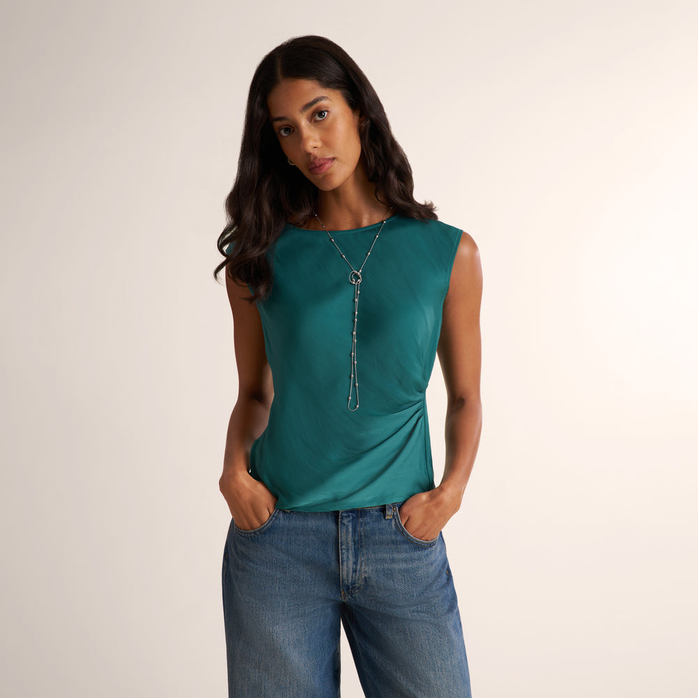 A woman modeling a sleeveless teal green top with a draped, gathered side detail, paired with blue jeans and a silver necklace, against a plain light beige background.
