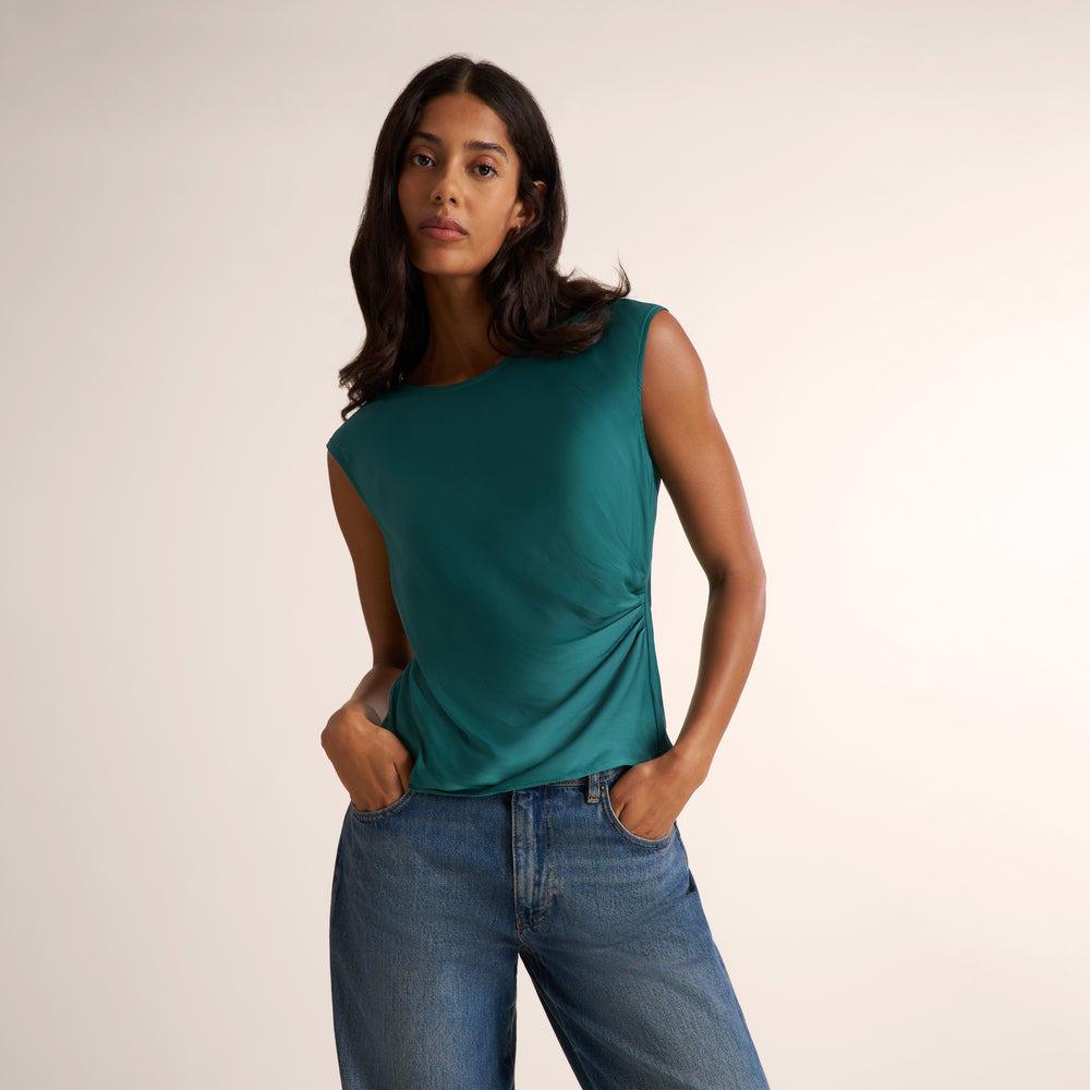 A woman modeling a sleeveless teal top with a draped side detail, paired with blue jeans, against a plain light beige studio background.