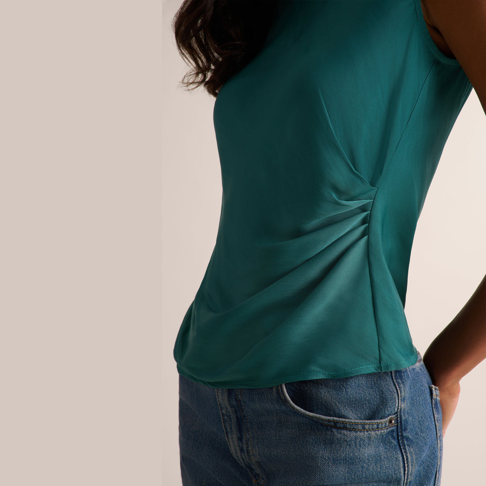 A close-up of a woman wearing a teal green sleeveless top with a gathered side detail, paired with blue jeans, against a plain light beige background.