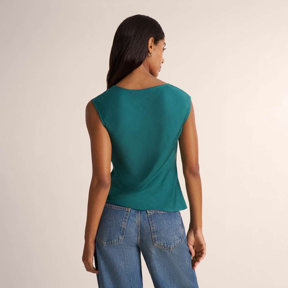 A woman shown from the back wearing a teal sleeveless top and blue jeans against a plain light beige studio background.