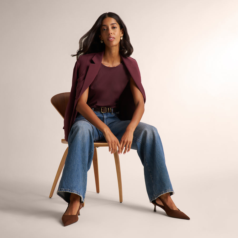 A woman sits on a wooden chair wearing a burgundy sleeveless top and matching draped blazer, blue wide-leg jeans, and brown pointed-toe heels against a plain light beige studio background.