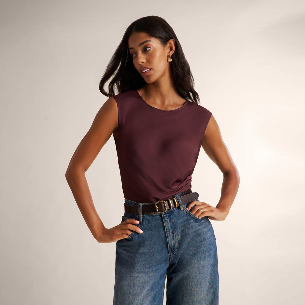 A woman modeling a sleeveless crew-neck top in deep burgundy, paired with blue jeans and a black belt, against a plain light beige studio background.