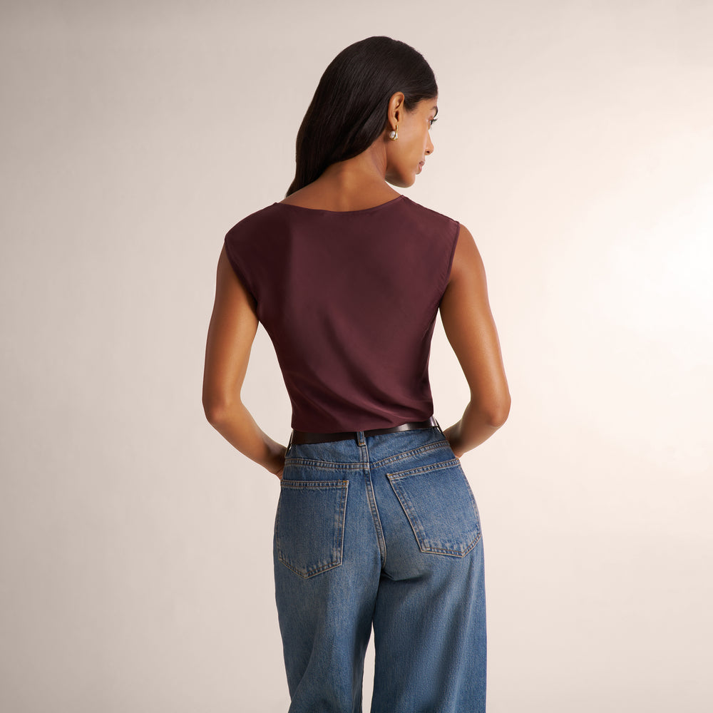 A woman shown from the back wearing a sleeveless deep burgundy top and blue jeans against a plain light beige studio background.