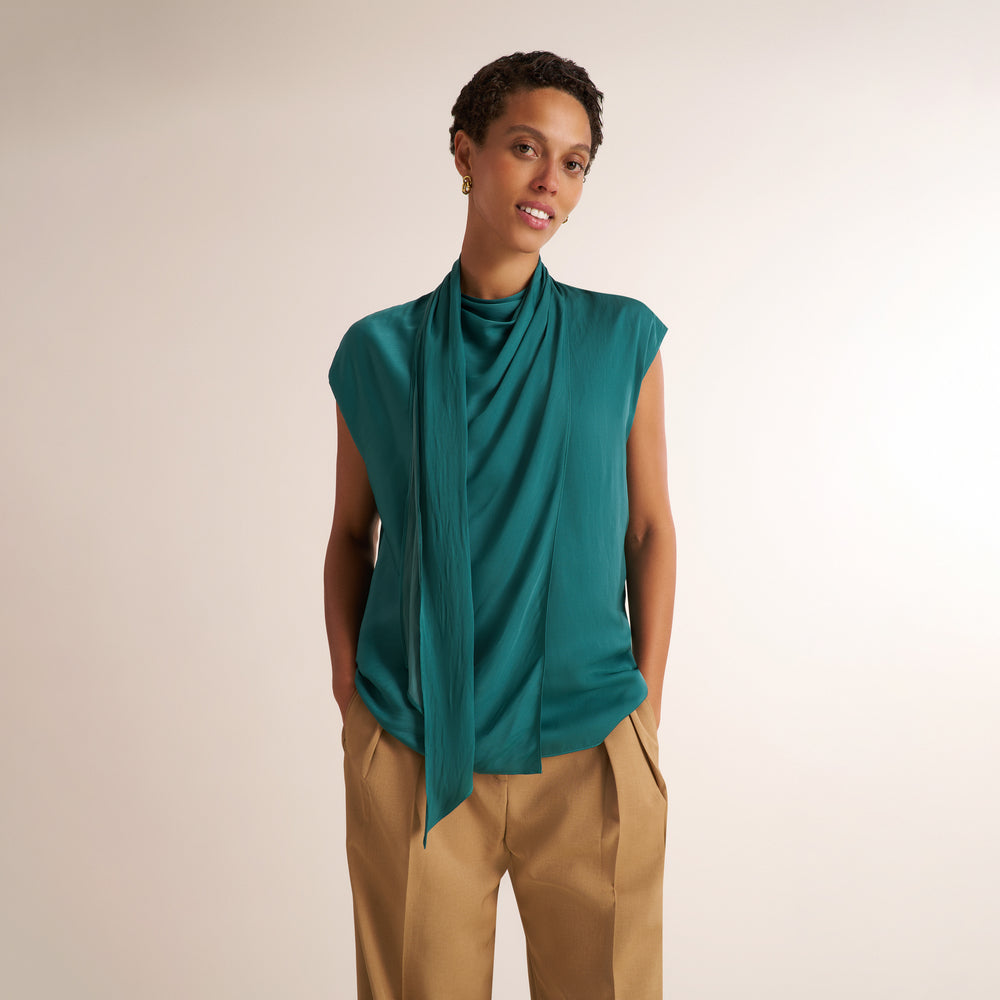 A woman modeling a teal green sleeveless draped top with a matching neck drape, paired with tan trousers, against a plain light beige studio background.