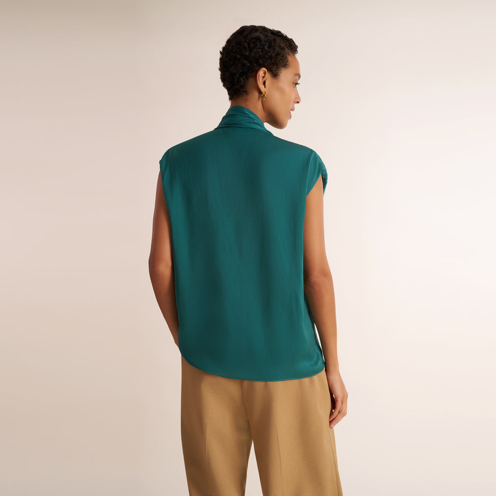 A model shown from the back wearing a sleeveless high-neck teal top and beige trousers, photographed against a plain light beige studio background.