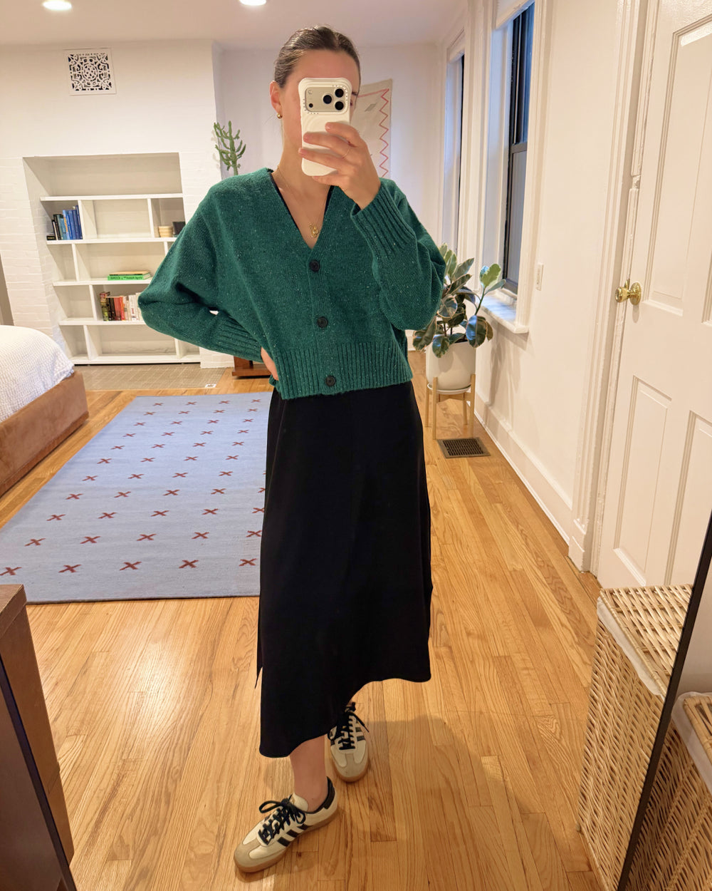 Person wearing a green cardigan and black skirt in a room with wooden flooring and white walls.