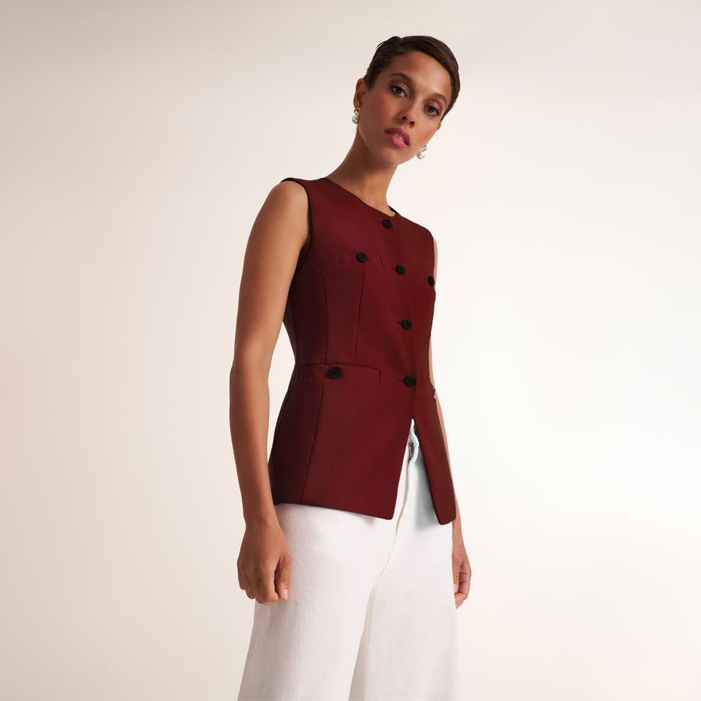 A woman modeling a sleeveless burgundy double-breasted vest with black buttons, paired with white trousers, against a plain light beige studio background.