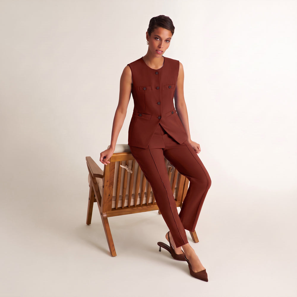 A model wearing a rust-brown tailored suit consisting of a sleeveless double-breasted vest and matching trousers, seated on a wooden chair against a plain off-white studio background.