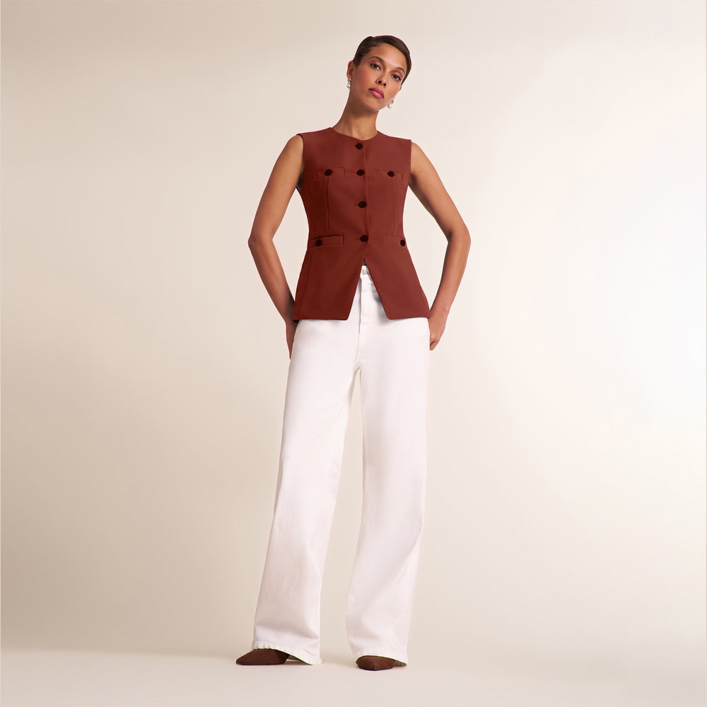 A model stands against a warm beige studio background wearing a sleeveless rust-brown vest with black buttons and white wide-leg trousers.