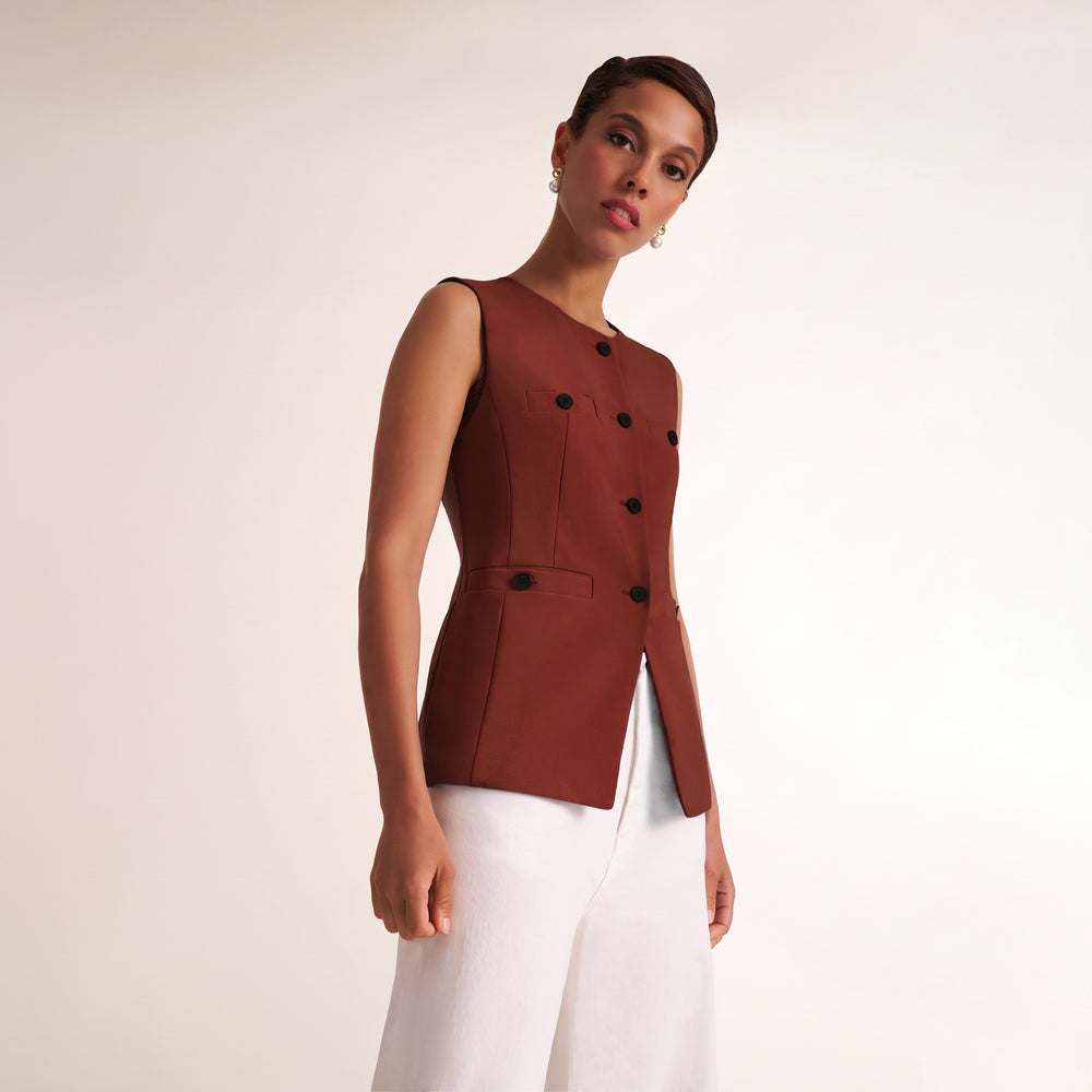A woman modeling a sleeveless burgundy double-breasted vest with black buttons, paired with white trousers, against a plain light beige studio background.