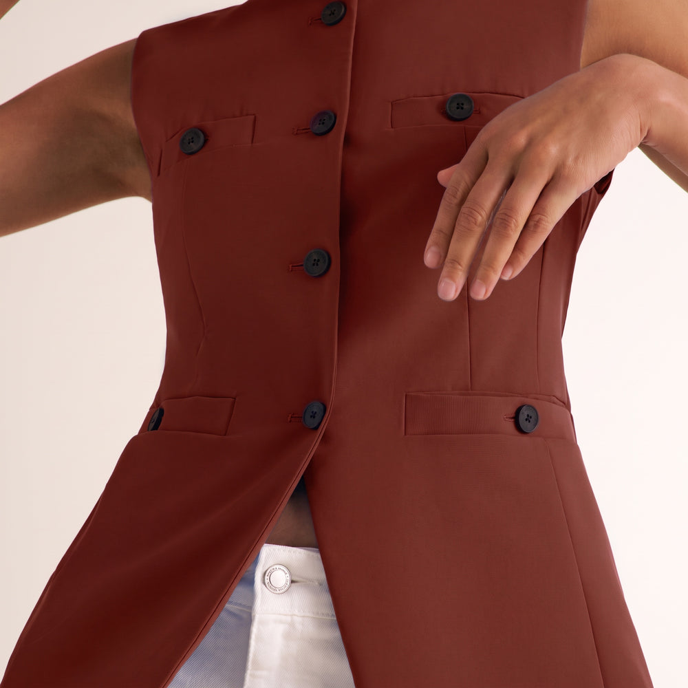 A close-up of a model wearing a sleeveless, structured burgundy vest with black buttons and flap pockets, paired with white pants against a light beige background.