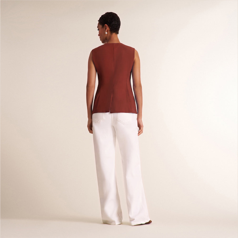 A model shown from the back wearing a sleeveless rust-brown structured top and white wide-leg trousers, standing against a plain cream studio background.