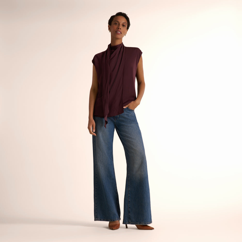 A model shown wearing a sleeveless, high-neck, dark burgundy top and blue jeans, standing against a plain light beige studio background.