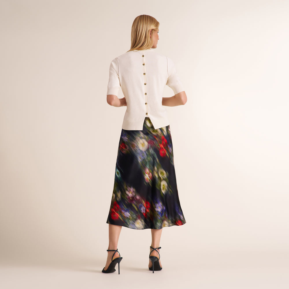 A woman shown from the back wearing a cream short-sleeve jacket with gold buttons and a dark midi skirt with a colorful floral print, paired with black high heels, against a plain beige studio background.