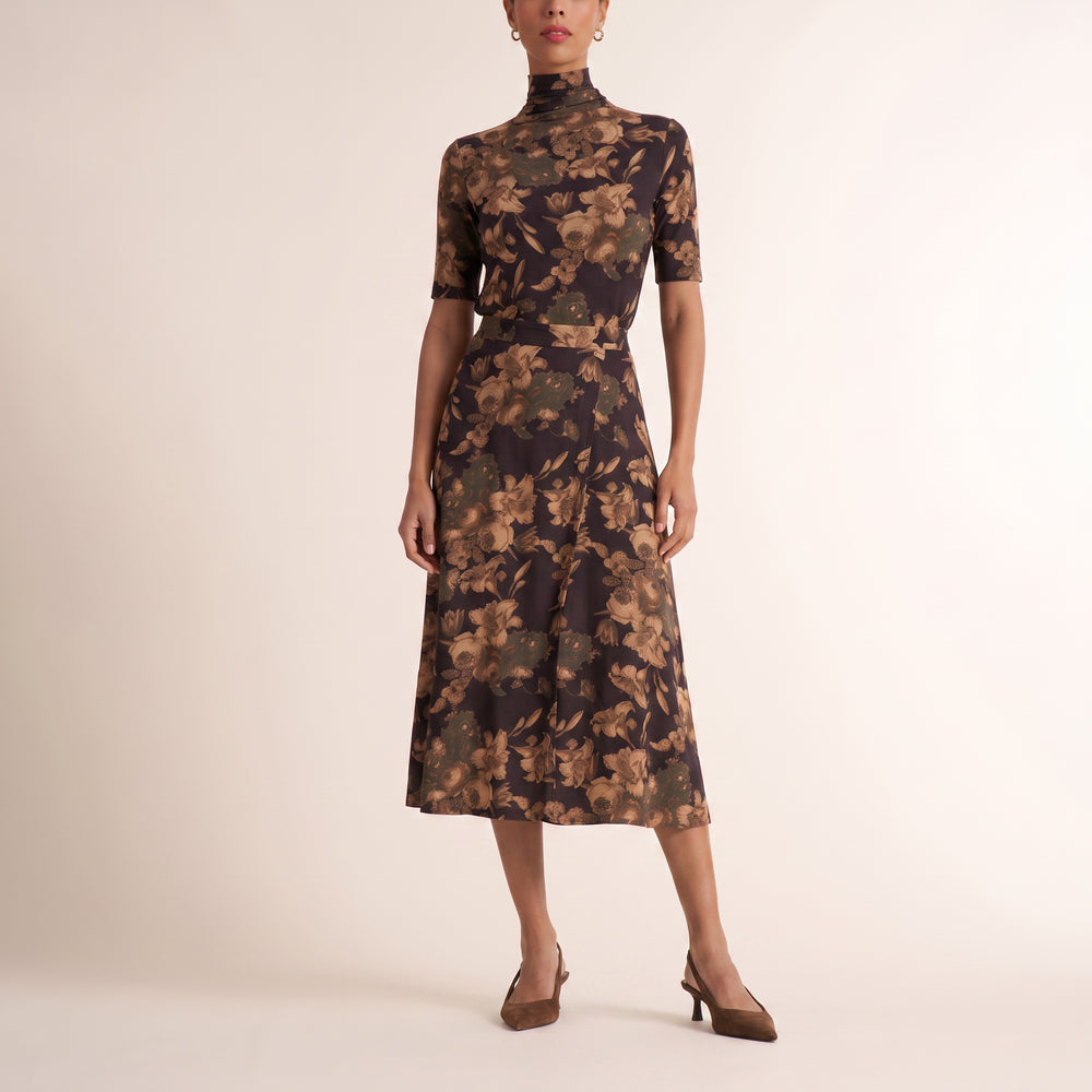 A model wearing a short-sleeved mock-neck midi dress with a brown and beige floral pattern on a dark background, paired with brown heels, against a plain beige studio backdrop.