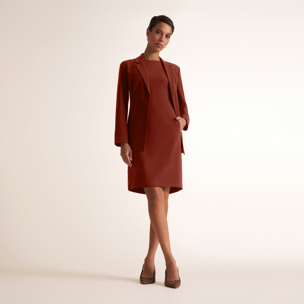 A woman modeling a rust-brown tailored blazer and matching knee-length dress against a plain light beige studio background.
