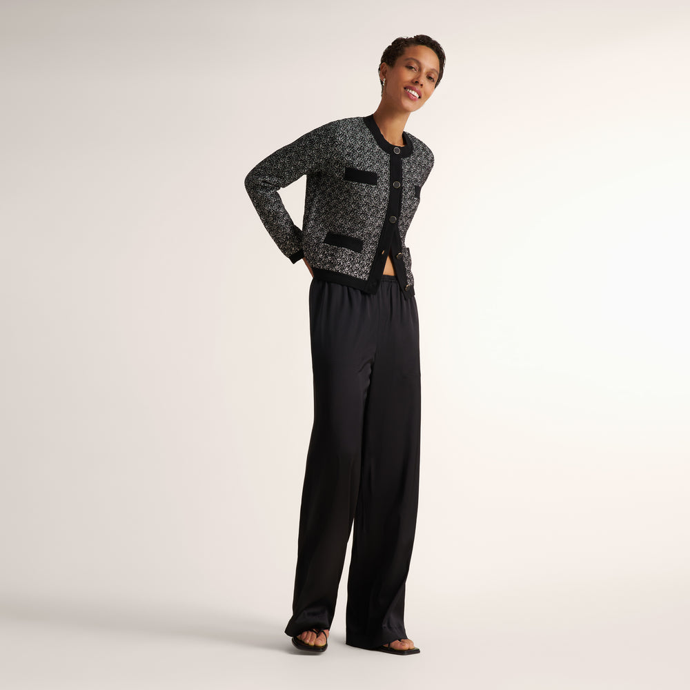 A woman modeling a black and gray patterned cropped cardigan with black trim, paired with black wide-leg trousers and black sandals, against a plain light gray studio background.
