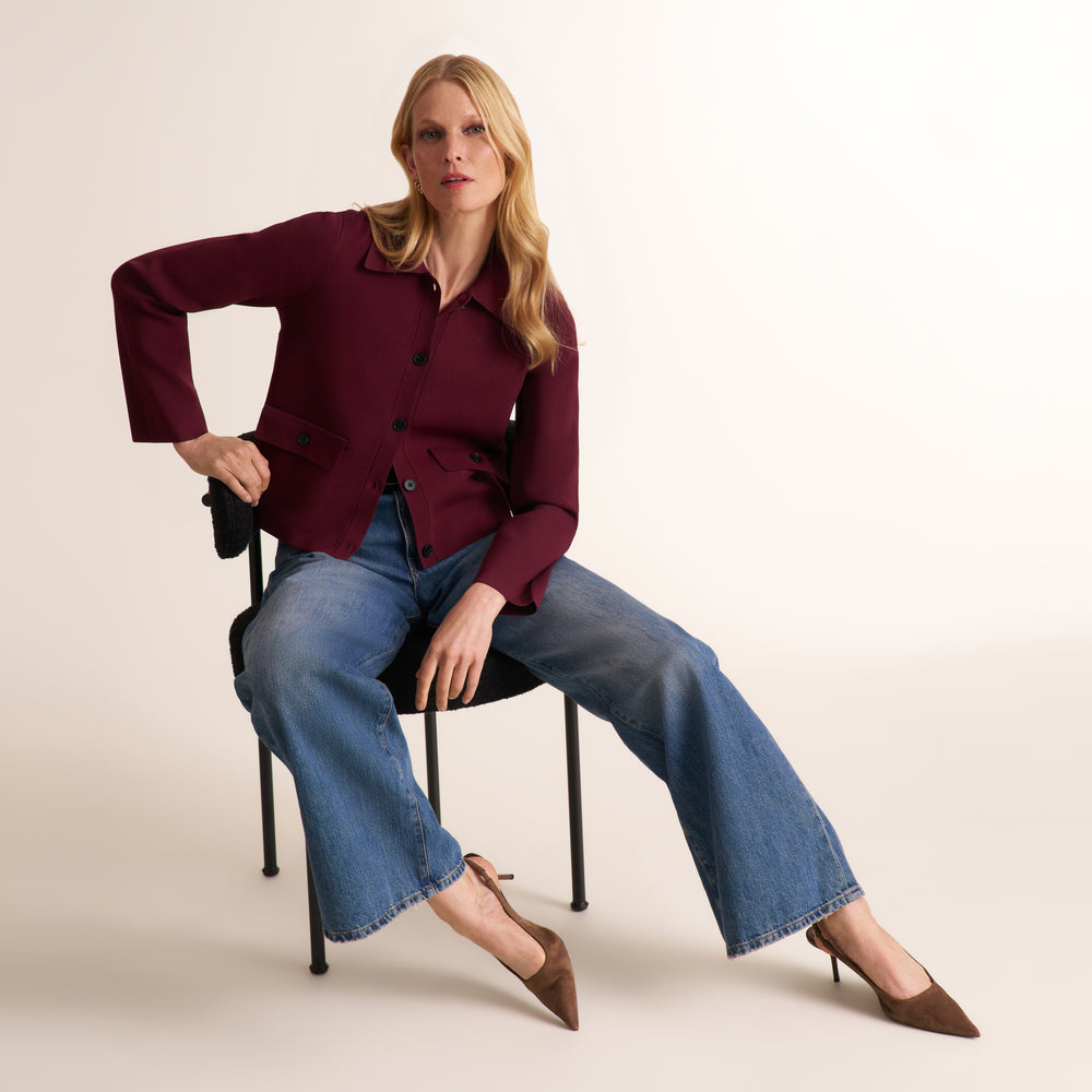 A woman sits on a black chair wearing a burgundy button-front jacket, wide-leg blue jeans, and brown pointed-toe heels against a plain light beige studio background.