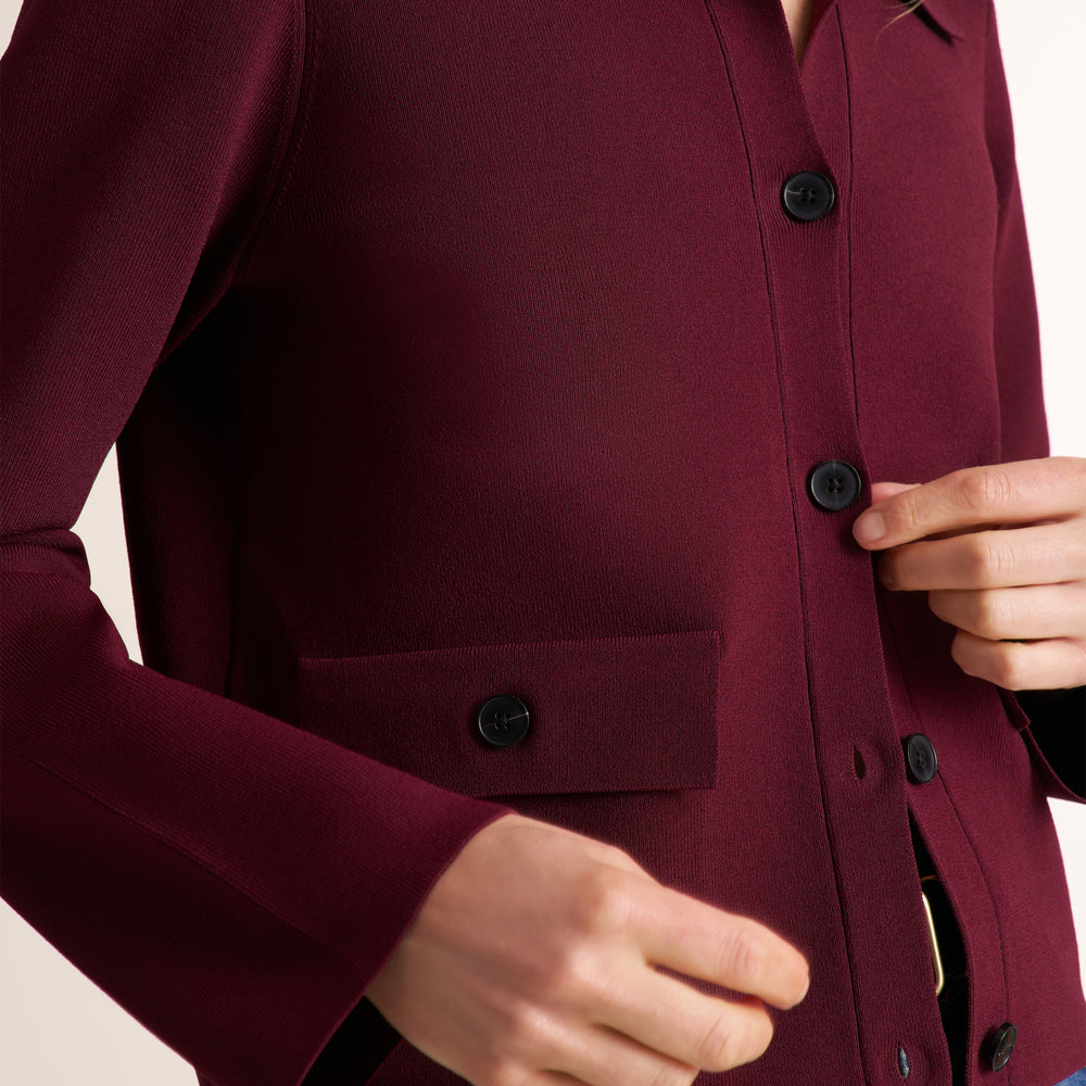 Close-up of a burgundy button-front cardigan or coat with a flap pocket, shown on a model with hands adjusting the front and pulling buttons.