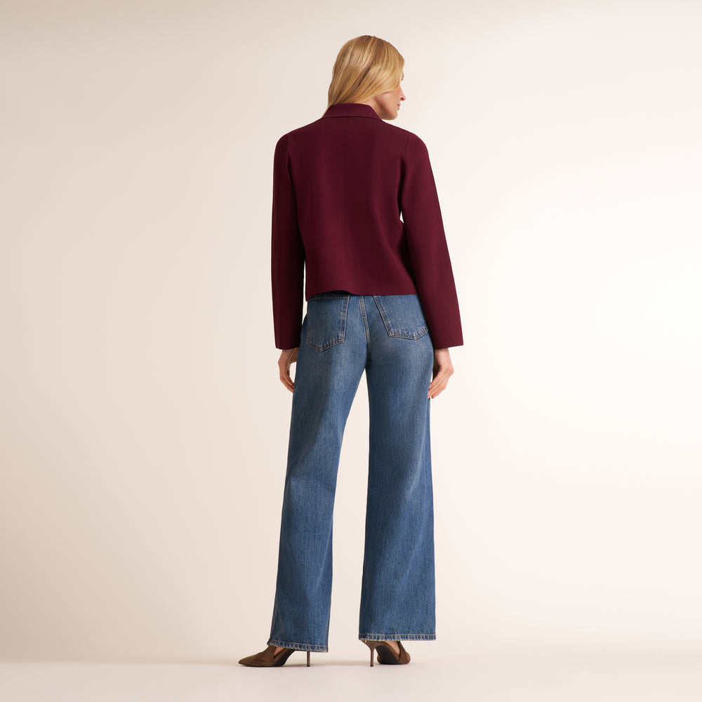 A woman is shown from behind wearing a cropped, long-sleeve burgundy knit top and wide-leg blue jeans, standing against a plain off-white studio background.