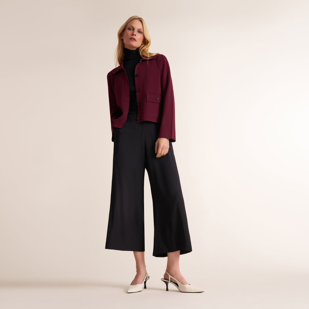 A woman models a dark burgundy cropped jacket over a black turtleneck, paired with black wide-leg cropped trousers and white slingback heels, against a solid light beige studio background.