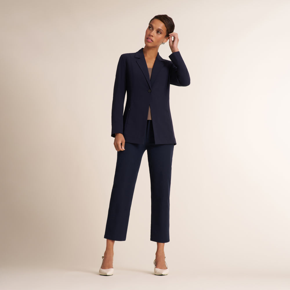 A woman modeling a navy blue two-piece suit consisting of a single-breasted blazer and matching cropped trousers, paired with white heels, against a solid light beige studio background.