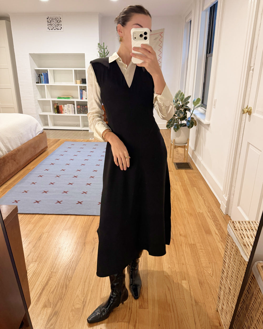 Person taking a mirror selfie wearing a black dress and white shirt in a room with wooden flooring and a bookshelf.