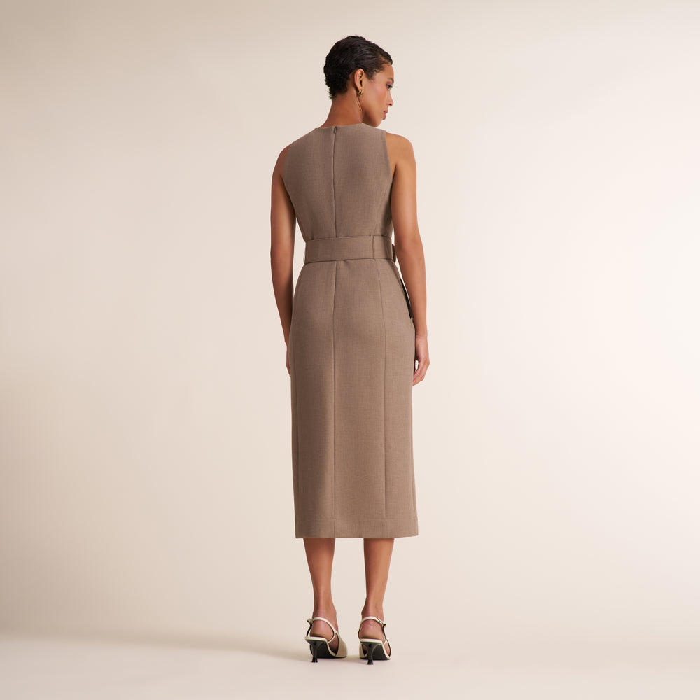 A woman stands with her back to the camera wearing a sleeveless, knee-length taupe dress with a defined waist and structured shoulders, paired with black and white heeled sandals, against a solid light beige studio background.